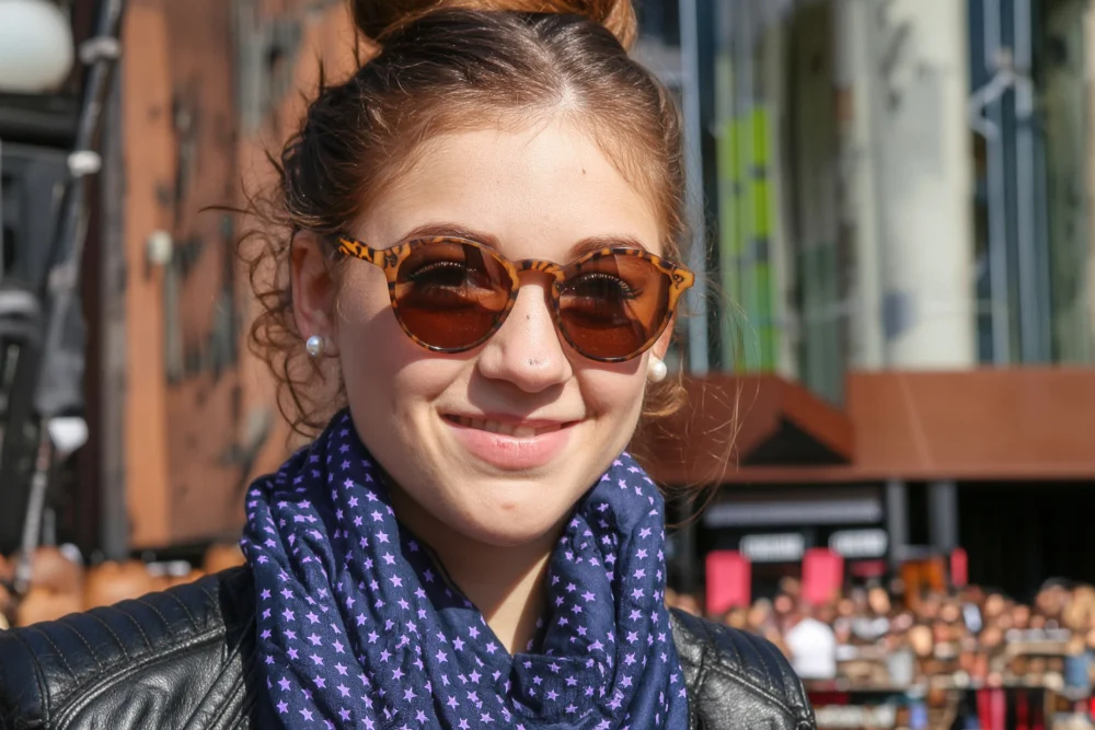 Generate an image that matches the exquisite silhouette outlined in Figure 1, and follows the following description: A young woman smiling on a sunny day, wearing a pair of brown round sunglasses with leopard print patterns on the frames. Her hair is neatly tied up, she wears pearl earrings, a dark blue scarf with purple star patterns around her neck, and a black leather jacket.