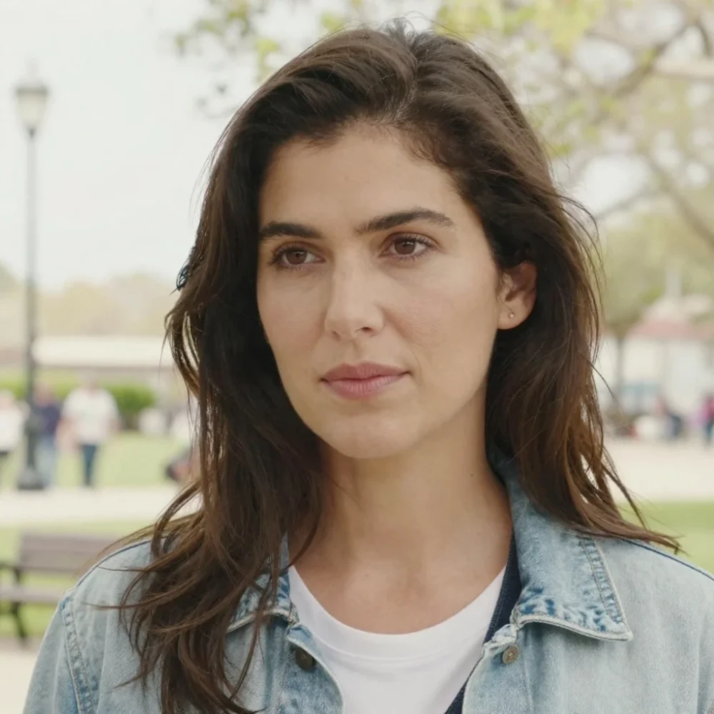 A realistic, high-resolution photograph of an American woman, captured in natural daylight with soft, diffused lighting that enhances her features gently. She is positioned in a candid, relaxed pose—standing outdoors near a park bench, wearing casual, contemporary clothing: a light blue denim jacket, a white t-shirt, and dark jeans. Her long, wavy brown hair falls loosely over her shoulders, and she has a subtle, thoughtful expression as she looks slightly off-camera. The background is softly blurred (shallow depth of field), with hints of greenery, a distant streetlamp, and a few passing pedestrians. Shot on a full-frame DSLR with a 50mm prime lens, emphasizing natural textures, skin tones, and environmental detail. Cinematic composition, eye-level shot, natural color grading, and a subtle film grain texture for authentic photographic realism.