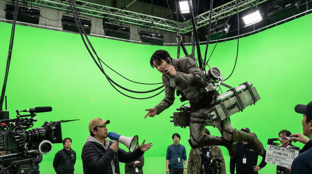 On-set documentary photo of filming a flying sequence. The actor playing Levi Ackerman is hanging upside down mid-air, suspended by a complex wire rigging harness against a gigantic green screen wall. He looks bored and is talking to the director down below. His ODM gear looks heavy, metallic, and weathered, with gas tanks and spare blades. Thick cables are visible attached to his waist. Industrial film lighting.