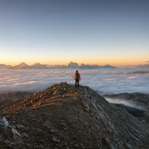 A wide landscape photograph of a lone hiker standing on a ridge above a sea of clouds at sunrise, golden light on distant snow-capped peaks, crisp detail, natural colors.