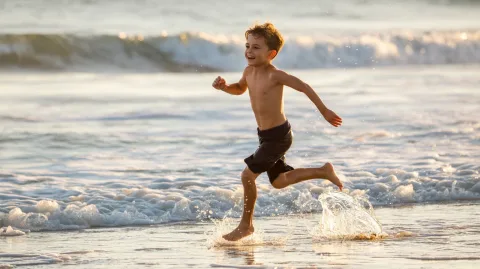 A cheerful boy runs barefoot across the beach, splashing through the shallow waves. The sun is bright, casting long shadows. The camera shakes slightly with his steps, enhancing the energy and joy of the scene.