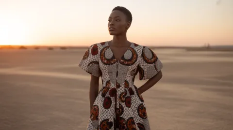Modern African woman in bold patterned Ankara dress, standing in a desert with a sunset backdrop, fashion editorial style, rich textures