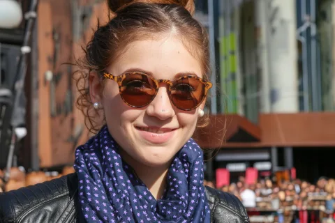 Generate an image that matches the exquisite silhouette outlined in Figure 1, and follows the following description: A young woman smiling on a sunny day, wearing a pair of brown round sunglasses with leopard print patterns on the frames. Her hair is neatly tied up, she wears pearl earrings, a dark blue scarf with purple star patterns around her neck, and a black leather jacket.