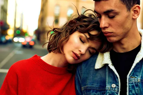 A european woman with short, tousled hair leans in close to a man, her eyes gently closed. She wears a glowing red sweater; he has a jeans jacket with a bright collar. Golden ambient light softens their skin tones. Their faces are calm and close, framed tightly in an intimate, cinematic shot. The background is a gentle blur of city motion, adding contrast to their stillness. Subtle film grain evokes a timeless, romantic feel.