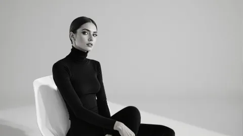 A minimalist fashion portrait of a woman in a sleek black turtleneck, seated on a modern white chair in an all-white room. Her gaze drifts off-camera, creating a cool, detached elegance. The composition is clean, with strong contrast and subtle shadow work.