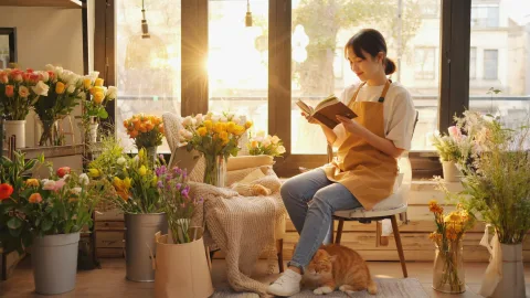 Inside a cozy, sunlit flower shop, a young female owner is focused on trimming a fresh bouquet of roses. She wears a linen apron, her hair casually tied up, with a gentle smile on her face. She is surrounded by buckets of various flowers, green plants, and rolls of wrapping paper, creating a scene of organized chaos. Soft morning light streams through the large street-front window, making the texture and colors of the petals incredibly vibrant.





In a cozy living room corner, a young woman is curled up in a soft armchair under a knitted blanket, peacefully reading a book. Warm afternoon sunlight streams generously through a large window, creating bright patches of light on the floor and visibly illuminating tiny dust motes dancing in the air. An orange tabby cat is curled up asleep by her feet. The entire scene exudes serenity, comfort, and a sense of peaceful contentment.




