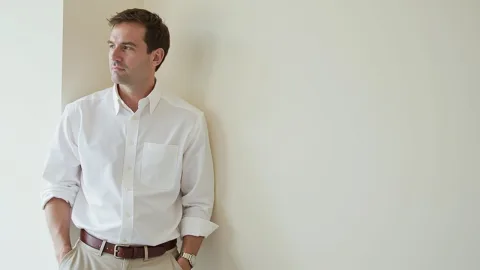 a man, leaning against a plain wall, one hand in his pocket, relaxed posture
