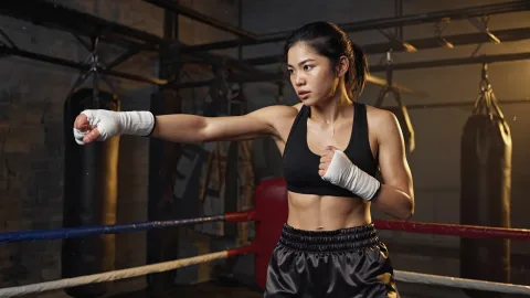 Action photograph of a female boxer during an intense training session in a gritty, dimly lit gym. She has just thrown a powerful right hook, and beads of sweat are flying off her brow. Her eyes show fierce determination, muscles tense and defined. The background is blurred with punching bags and a boxing ring. Dramatic side lighting highlights the contour of her body and the flying sweat droplets.



