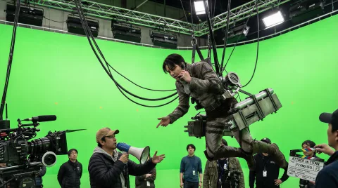 On-set documentary photo of filming a flying sequence. The actor playing Levi Ackerman is hanging upside down mid-air, suspended by a complex wire rigging harness against a gigantic green screen wall. He looks bored and is talking to the director down below. His ODM gear looks heavy, metallic, and weathered, with gas tanks and spare blades. Thick cables are visible attached to his waist. Industrial film lighting.