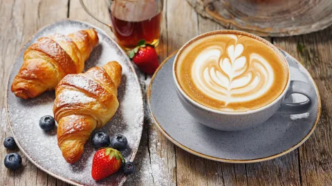 Food photography of a delicious brunch set on a rustic wooden table next to a window with soft morning light. On the plate are two golden-brown croissants with powdered sugar, next to a few fresh strawberries and blueberries. A steaming cup of latte with perfect latte art. macro shot, extremely detailed.

